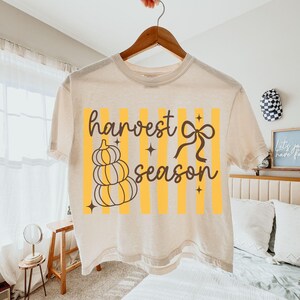 May include: Cream-colored t-shirt with a fall-themed graphic. The design features a stack of pumpkins, a bow, and the words "harvest season" in a stylish font, set against yellow vertical stripes.