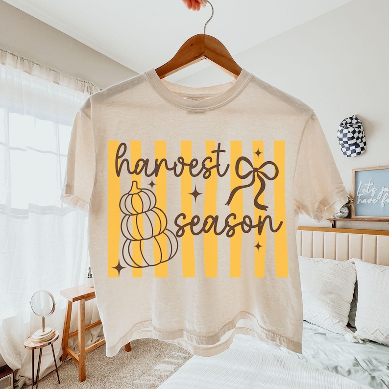 May include: Cream-colored t-shirt with a fall-themed graphic. The design features a stack of pumpkins, a bow, and the words "harvest season" in a stylish font, set against yellow vertical stripes.