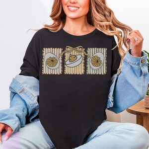 May include: Black sweatshirt featuring a graphic of three deviled eggs in decorative frames. The eggs have a tan filling and are set against a striped background with a bow. A casual, comfortable top for Easter.
