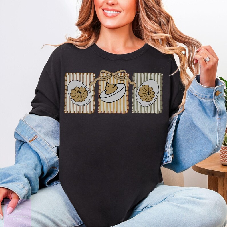 May include: Black sweatshirt featuring a graphic of three deviled eggs in decorative frames. The eggs have a tan filling and are set against a striped background with a bow. A casual, comfortable top for Easter.