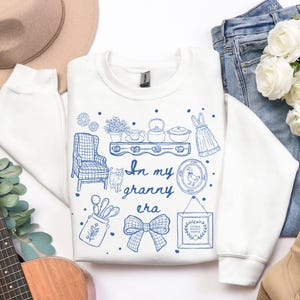 May include: White sweatshirt with a blue illustration of a shelf with teacups, a teapot, and a dress. The text "In my granny era" is written in blue script. Other illustrations include a chair, a cat, and a framed picture.