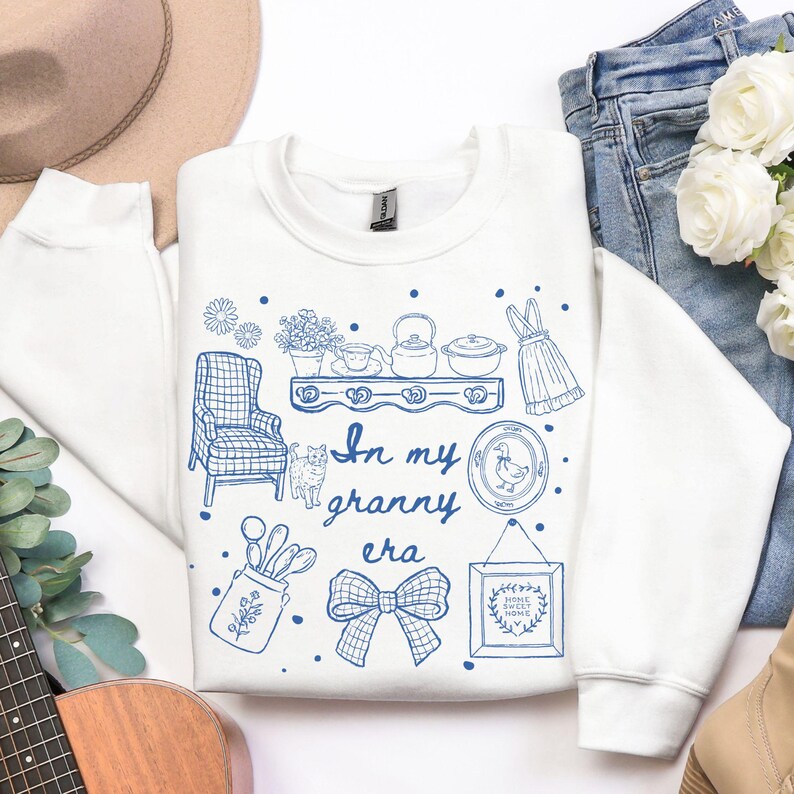 May include: White sweatshirt with a blue illustration of a shelf with teacups, a teapot, and a dress. The text "In my granny era" is written in blue script. Other illustrations include a chair, a cat, and a framed picture.