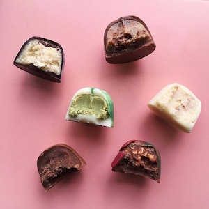 May include: Six different chocolate truffles with various fillings, including green tea, vanilla, and chocolate. The truffles are arranged on a pink surface.