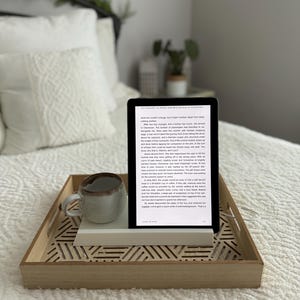 May include: A wooden tray with a geometric pattern holds a white e-reader, a white ceramic mug, and a book. The e-reader is displaying a page of text from a novel. The tray is on a white bedspread.