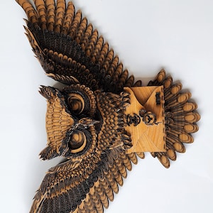 May include: A wooden owl wall decor with intricate laser cut details. The owl has outstretched wings and a detailed face with large eyes. The owl is holding a small wooden envelope in its talons.