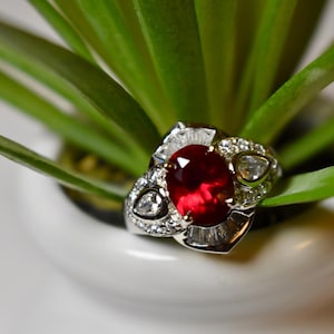 Könnte beinhalten: Ein silberner Ring mit einem großen, ovalen Rubin-Edelstein, der von kleineren Diamanten umgeben ist. Der Ring ist in einem filigranen, komplizierten Design gefasst.
