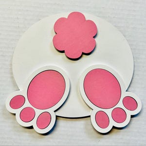 May include: A wooden Easter decoration featuring a white circular base with a pink bunny tail and two pink bunny feet. The bunny tail is a pink flower shape. The bunny feet have pink pads and white outlines.