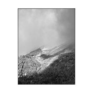May include: Black and white framed print of a mountain range. The image captures a snow-covered mountain with a dense forest at its base. The sky is overcast, creating a dramatic contrast between light and shadow.