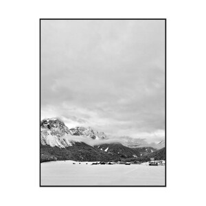 May include: Black and white landscape photograph of a mountain range covered in snow, with a cloudy sky overhead. The image features a valley with small buildings at the base of the mountains, creating a serene and natural scene.