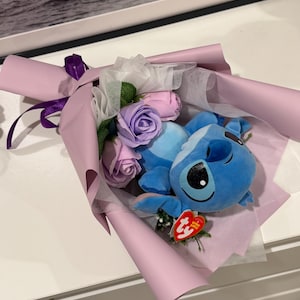 May include: A bouquet featuring a blue plush character with large black eyes, nestled among pink and purple artificial roses. The arrangement is wrapped in light purple paper and tied with a purple ribbon. A red tag is attached to the plush toy.
