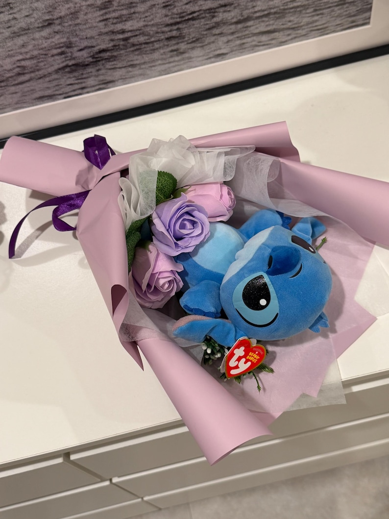 May include: A bouquet featuring a blue plush character with large black eyes, nestled among pink and purple artificial roses. The arrangement is wrapped in light purple paper and tied with a purple ribbon. A red tag is attached to the plush toy.