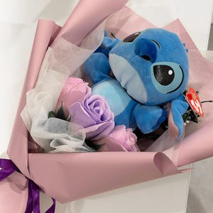 May include: A bouquet featuring a blue plush toy character nestled among pink and lavender artificial roses. The arrangement is wrapped in pink paper and tied with a purple ribbon, creating a whimsical gift.