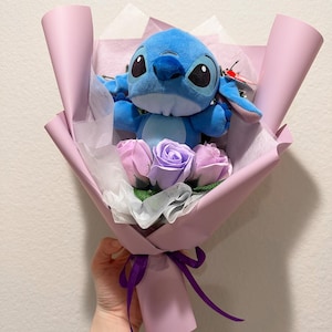May include: A bouquet featuring a blue plush character with large black eyes, nestled among lavender and pink roses. The arrangement is wrapped in pink paper and tied with a purple ribbon, creating a whimsical and charming gift.
