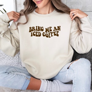 Könnte beinhalten: Ein beiger Pullover mit dem Text "Bring me an iced coffee" in braunen Retro-Lettern.