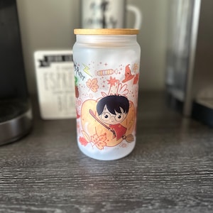 Puede incluir: Un tarro de vidrio esmerilado con tapa de madera con un diseño temático de Harry Potter. El tarro tiene una ilustración colorida de Harry Potter volando en una escoba con una calabaza y hojas de otoño. El texto "Harry Potter" es visible en el tarro.