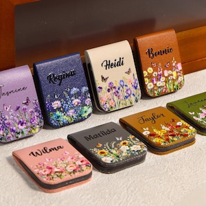 Puede incluir: Una colección de pinzas para billetes magnéticas personalizadas de cuero. Cada pinza presenta un diseño floral con mariposas y un nombre en letra cursiva. Colores: morado, azul, beige, marrón, rosa, gris y verde.