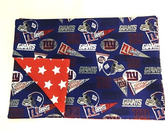 May include: A reversible placemat in blue and red with a pattern of New York Giants logos and stars. The front side features a blue background with white and red New York Giants logos and the back side features a red background with white stars.