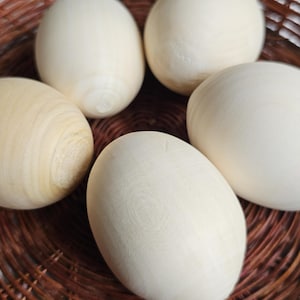 Può includere: Cinque uova di legno lisce e di colore chiaro sono in un cestino marrone intrecciato. Le uova hanno una finitura naturale, non verniciata, che mostra la venatura del legno. La trama del cestino contrasta con le superfici lisce delle uova.