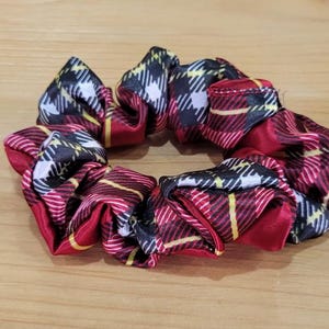 Può includere: Un elastico per capelli a quadri rossi, neri e bianchi. Il tessuto ha una lucentezza satinata ed è raccolto per creare una forma circolare voluminosa. Linee gialle sono visibili nel motivo a quadri. L'elastico è un accessorio per capelli.