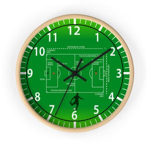 Puede incluir: Un reloj de pared redondo con marco de madera clara. La esfera verde muestra un diseño de campo de fútbol con números y manecillas blancas. El reloj usa manecillas negras para indicar la hora.