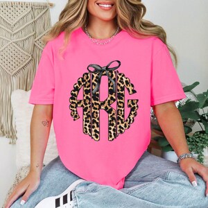 May include: A pink t-shirt with a black bow and a leopard print monogram. The monogram is in the shape of a circle and has the letters "ARG" in it.