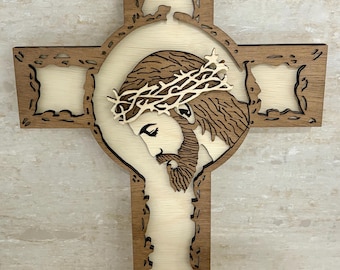 May include: Wooden cross featuring a detailed depiction of Jesus wearing a crown of thorns. The cross has a layered design with a light-colored background and a dark brown frame. The image of Jesus is intricately carved.