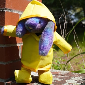 May include: A plush bunny toy with purple and blue fur is wearing a bright yellow raincoat and matching boots. The raincoat has a hood and orange trim. The bunny is standing next to a red brick wall.