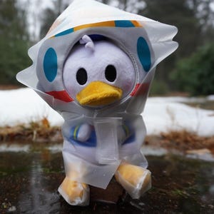 Könnte beinhalten: Ein weißer Plüsch-Enten mit einem blau-weißen Astronautenkostüm und einem durchsichtigen Plastik-Regenmantel. Die Ente sitzt auf einer nassen Oberfläche mit Schnee im Hintergrund.
