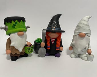 May include: Three Halloween-themed gnome figurines. The first is a Frankenstein monster gnome with a green head, bolts, and a candy bucket. The second is a witch gnome with a black hat, red braids, and a cauldron. The third is a white mummy gnome.