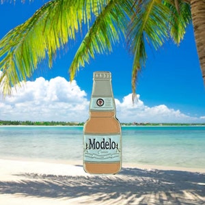 May include: A gold-coloured enamel pin of a brown glass bottle with a white label that says "Modelo" on it. The bottle is in front of a beach scene with a palm tree and blue sky.