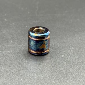 May include: A small, cylindrical bead with a hole through the center. The bead has a metallic finish with blue, gold, and purple hues. The top and bottom edges are outlined in gold. The bead is likely used for jewelry making or crafts.