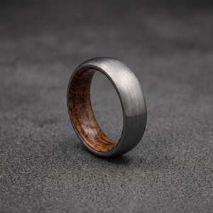 Könnte beinhalten: Ein Ring mit einem gebürsteten grauen Metallaußenband und einer Einlage aus warmem braunem Holz mit sichtbarer Maserung auf der Innenseite. Der Ring hat ein glattes, abgerundetes Profil mit einer sichtbaren Textur auf der Außenseite.