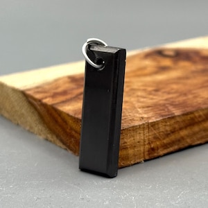 May include: A black rectangular pendant with a silver-colored loop at the top. The pendant is displayed on a wooden surface with a natural grain pattern. The background is a neutral gray.