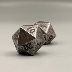 May include: Two metal D20 dice with a wood grain pattern. The dice are silver with black numbering. One die shows the number 10, and the other die is partially visible. The dice are for tabletop games.