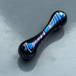May include: A small, elongated, decorative object with a blue, black, and silver swirled pattern. The object has a rounded shape at both ends and a narrower center. The surface reflects light, creating a metallic sheen.