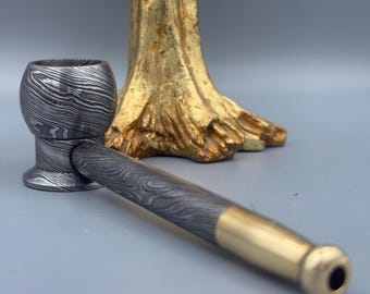 May include: A handcrafted pipe with a patterned bowl and stem, featuring a brass end. The pipe has a unique, textured design and is set against a blurred background with a gold-colored decorative element.