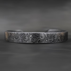Puede incluir: Una pulsera de plata con un patrón arremolinado único. La banda de metal tiene una superficie texturizada con una apariencia oscura y moteada. El diseño abierto permite un uso fácil. Un accesorio elegante.