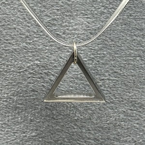 Puede incluir: Un collar con colgante plateado con un diseño de triángulo geométrico. El colgante cuelga de una fina cadena plateada. El triángulo tiene un centro hueco y una estética simple y moderna. El collar se muestra sobre un fondo gris.