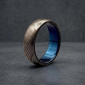 Pode incluir: Close-up de um anel com um exterior metálico texturizado e um interior azul vibrante. A superfície do anel tem um padrão de linhas diagonais, criando uma textura visual única. O anel é uma joia.