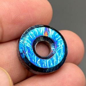 May include: A small, circular metal object with a central hole. The outer ring features a vibrant blue and purple lightning bolt pattern. The object has a dark, metallic finish and appears to be a decorative element or component.