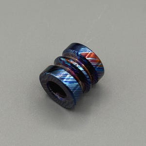 May include: A multi-colored bead with a layered design. The bead features a combination of blue, orange, and purple hues. The bead has a cylindrical shape with a hole running through the center.