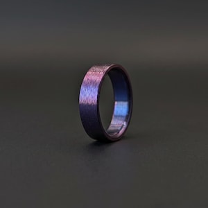 May include: A circular ring featuring a brushed metal texture with a gradient of iridescent colors, transitioning from deep violet and purple tones to shades of blue. The band has a smooth, polished interior surface and a textured exterior finish.