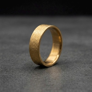 Könnte beinhalten: Ein goldfarbener Ring mit strukturierter Oberfläche. Der Ring hat ein schlichtes Banddesign, wahrscheinlich für eine Hochzeit oder einen Verlobungsring. Der Ring besteht aus einem glänzenden, metallischen Material.