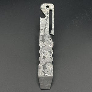May include: A Damascus steel multi-tool featuring a bottle opener, a slotted section, and a textured grip. The tool has a dark, swirling pattern and a rectangular base. A small hole is at the top for attachment.