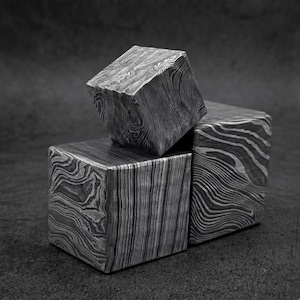 May include: Three solid metal cubes featuring a distinct Damascus steel pattern with swirling, layered lines in shades of gray, black, and silver. The cubes are arranged in a stacked formation, showcasing the intricate, flowing metallic grain.