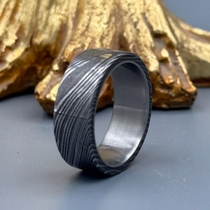 May include: A close-up of a Damascus steel ring. The ring has a dark, patterned exterior with a silver interior. The ring is set against a gold-colored, textured background and a gray surface.