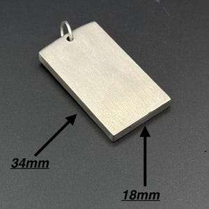 Könnte beinhalten: Ein rechteckiger silberner Anhänger mit einer Schlaufe für eine Kette. Der Anhänger ist 3,4 cm lang und 1,8 cm breit. Die Oberfläche hat ein gebürstetes Metall-Finish. Der Anhänger wird vor einem dunklen Hintergrund gezeigt.