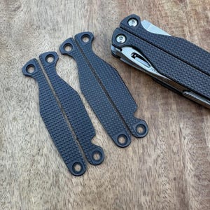Op de afbeelding: Een multitool en verschillende donkergrijze vervangende handgreepschalen zijn gerangschikt op een houten oppervlak. De schalen hebben een textuur met een rasterpatroon en ronde gaten. De multitool is gedeeltelijk geopend en toont een mes en andere gereedschappen.