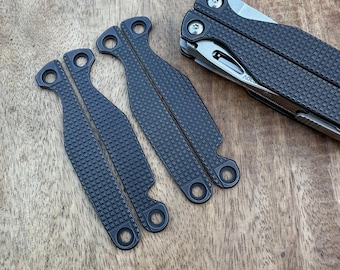 Könnte beinhalten: Ein Multitool und mehrere dunkelgraue Ersatzgriffschalen sind auf einer Holzoberfläche angeordnet. Die Schalen haben ein strukturiertes, gitterartiges Muster und kreisförmige Löcher. Das Multitool ist teilweise geöffnet und zeigt eine Klinge und andere Werkzeuge.