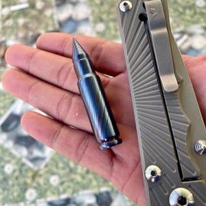 Może przedstawiać: Iryzująca łuska pocisku w kolorze niebieskim i fioletowym obok szarego noża kieszonkowego z napisem "IDAHO" na rękojeści.